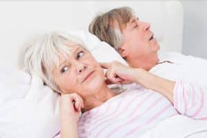 Do you or a loved one suffer from sleep apnea? Do you often wake up tired and groggy in the middle of the night because you have no control over your breathing? Image of a elderly couple in bed and the woman has her fingers in her ears to drown out the sound of the mans snoring.
