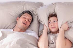 Oral Appliance from Lee Family Dentistry For Sleep Apnea is an affordable and effective solution for people experiencing mild to moderate obstructive sleep apnea. Image of a young couple in bed and the woman has a pillow over her ears to drown out the sound of the mans snoring.