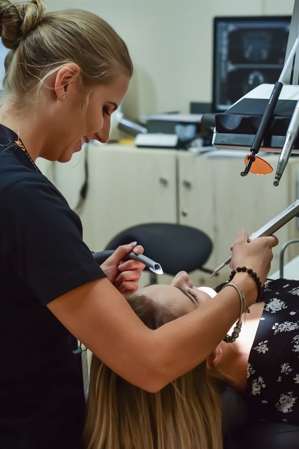 A smiling, blonde, dental hygienist leans over a happy patient who is in a dental chair.