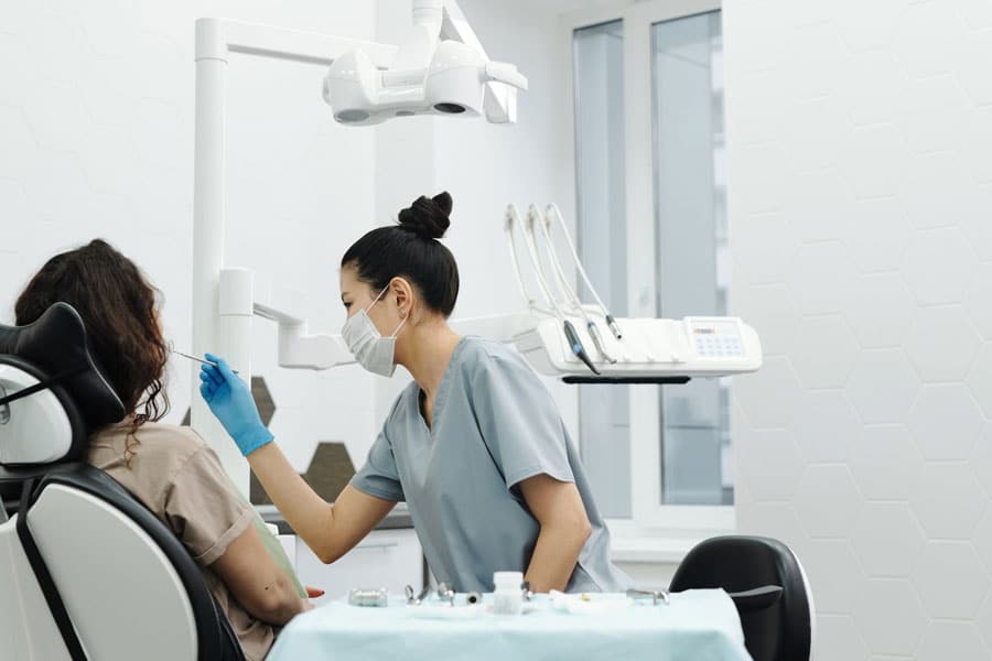A brunette woman sitting in a dentist's chair while a dark haired dental hygienist in gray scrubs and a mask and blue glove leans over her.